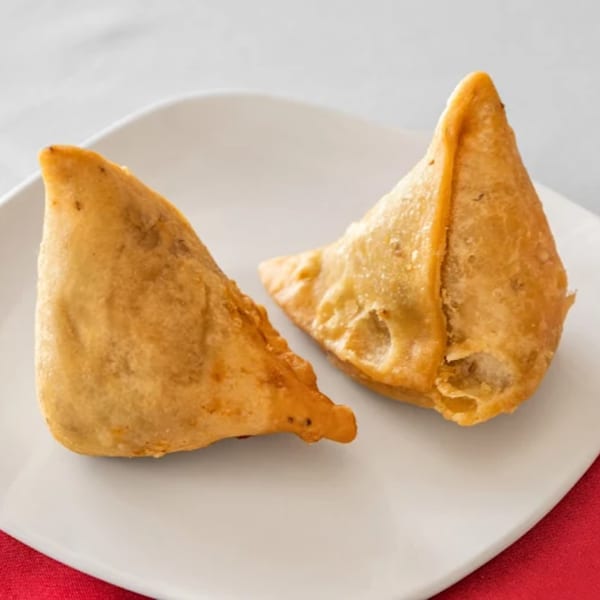 Two golden-brown, triangular samosas rest on a white plate with a red napkin underneath.