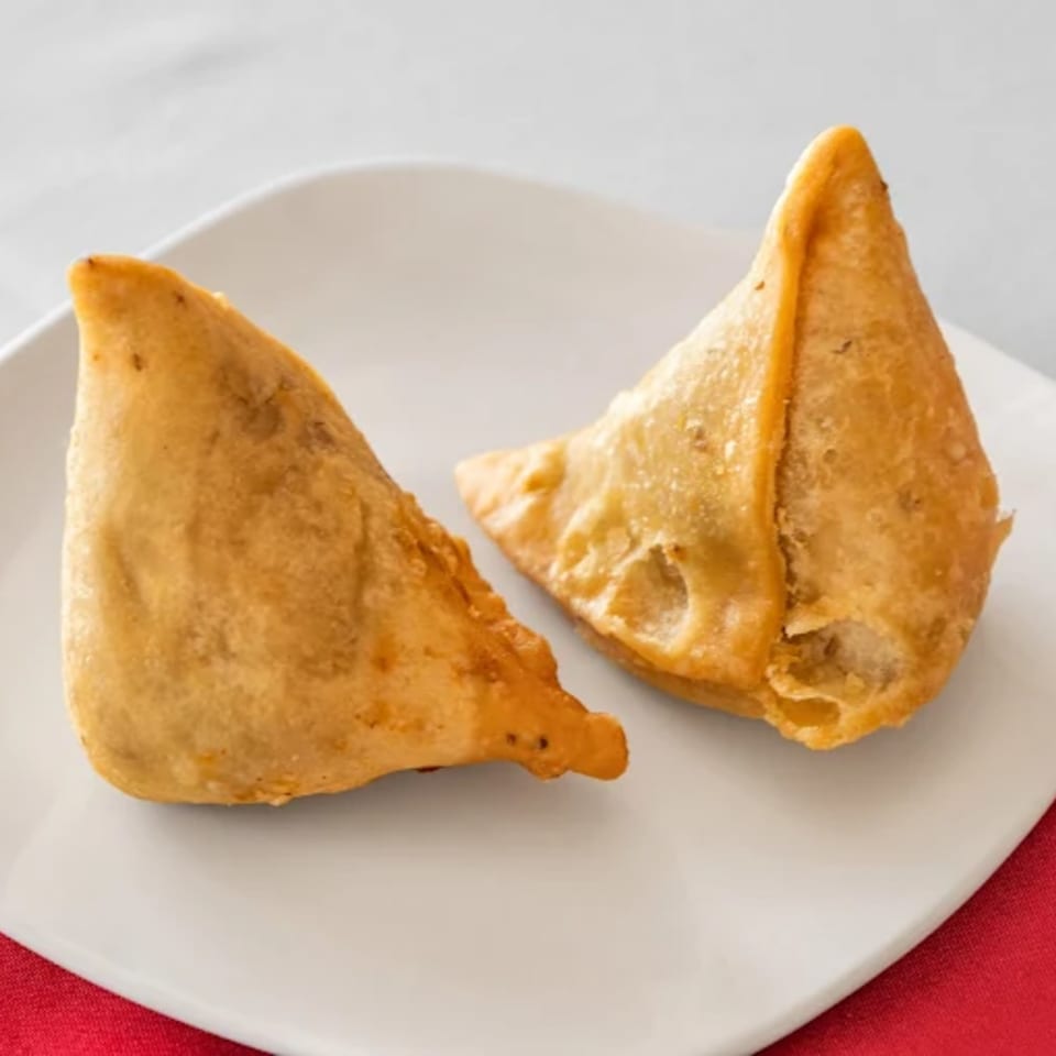 Two golden-brown, triangular samosas rest on a white plate with a red napkin underneath.