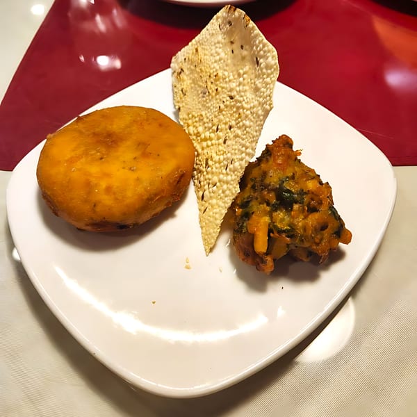 A white plate holds a golden fried pastry, a crispy papadum, and a vegetable fritter.